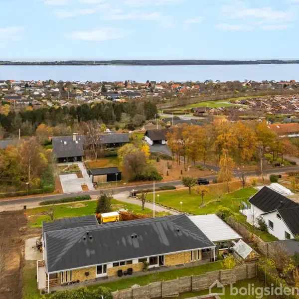 Hus ligger i et boligområde med grønne områder og udsigt til en sø. I forgrunden ses en vej og flere huse. Træer med efterårsløv omkranser området.