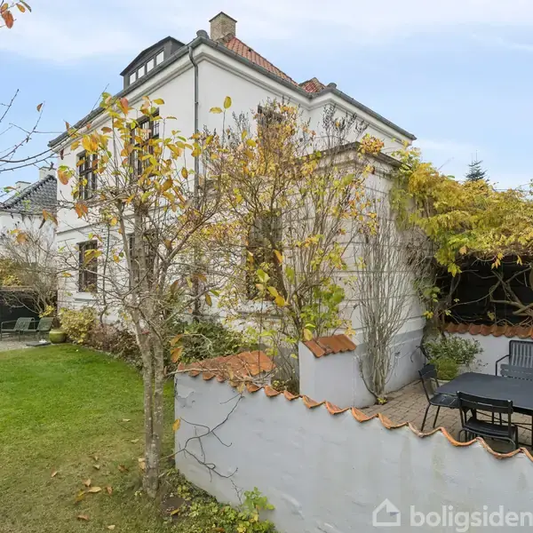 Et hvidt hus med to etager står blandt efterårsløv. Haven har en terrasse med et sort bord og stole, omgivet af grønne buske og træer under en klar himmel.