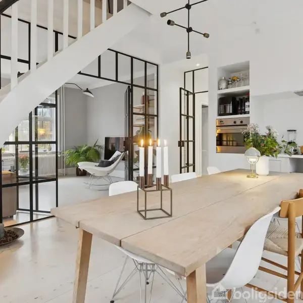 Et spisebord med stole er placeret under en elegant loftslampe i et lyst, moderne åbent køkken-alrum. En trappe fører opad, og planter giver liv til rummet.