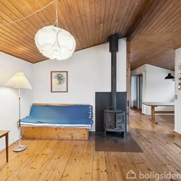 Brændeovn står i stue med trægulv og træloft, ved siden af en blå sofa og gulvlampe. Der er et maleri på væggen bagved, og et bord ses i baggrunden.