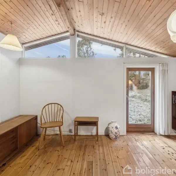 Et lyst værelse med trægulv og træloft. En træstol og et lille bord står ved siden af et hvidt vase. En reol fyldt med bøger står til højre.