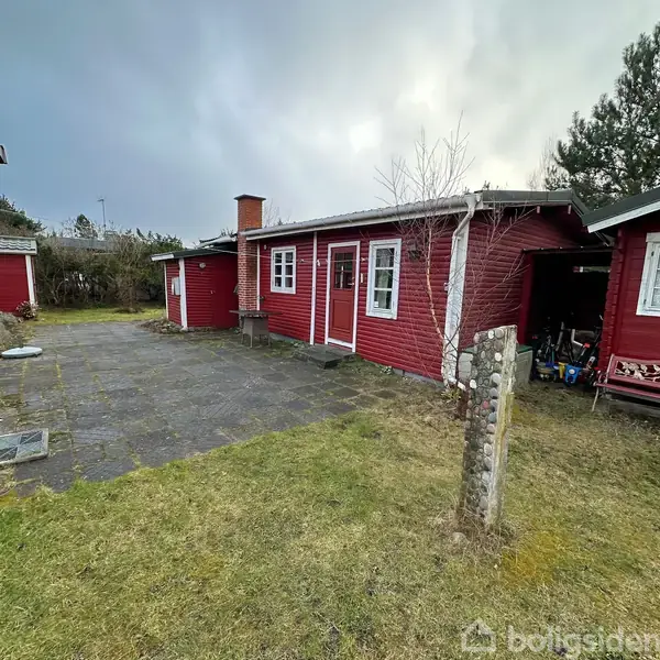 Rødt træhus med en skorsten, omkranset af fliser og græsplæne. Til højre ses en garage med åbent skur. Huset har to vinduer og en rød dør.