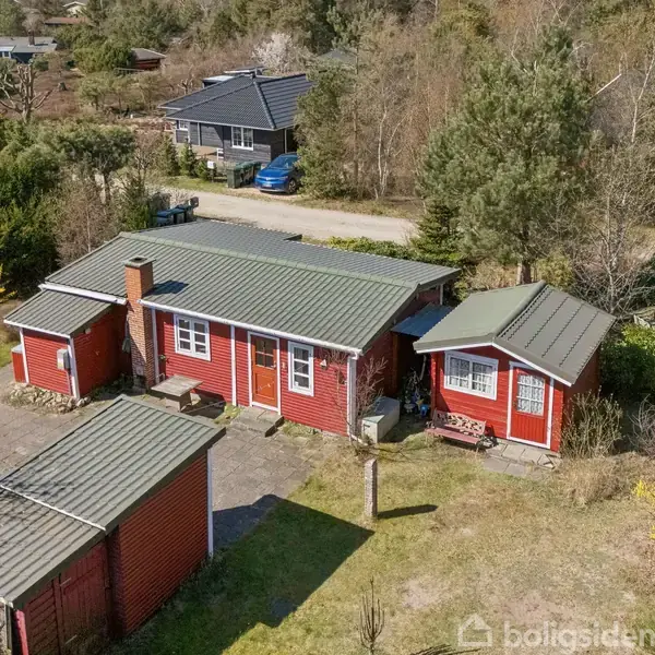 Rødt træhus med grønne tage står stille omgivet af træer og en beskeden have. I baggrunden ses flere huse samt en vej med en parkeret blå bil.
