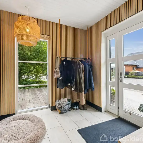 Entré med tøjstativ fyldt med jakker, placeret på flisegulv. Rundt tæppe ved glasdør leder til terrasse med udsigt til haven. Væggene har lodrette træpaneler.