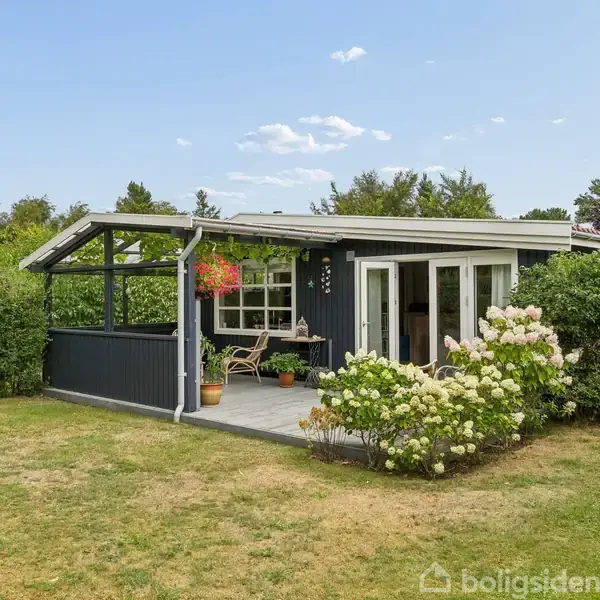 Hyggeligt sommerhus med mørke trævægge og hvidt tag; står stille på en velplejet græsplæne med blomsterbuske og havemøbler på terrassen; omgivet af grønne planter og blå himmel.