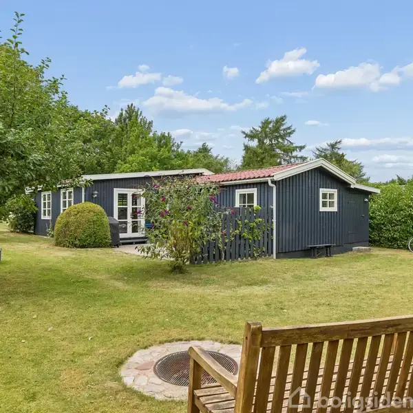Et sort sommerhus står i en grøn have med træer og buske. En bænk og cykler er placeret i nærheden. Skydækket himmel danner baggrunden.