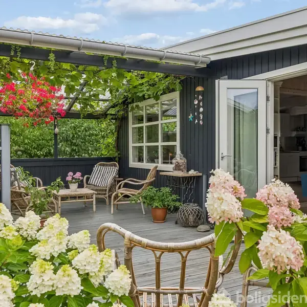 En terrasse med træmøbler og blomsterkrukker under en grøn pergola. Området er omgivet af frodig have med blomstrende planter, og husets åbne glasdøre fører ind til stuen.