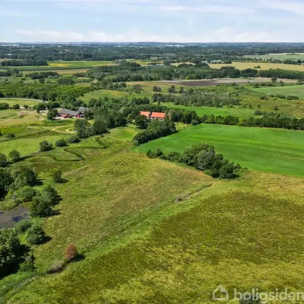 Landskab med grønne marker og træer strækker sig ud i horisonten under en blå himmel. En rød bygning ses omgivet af grønt. Placering: Boligsiden.