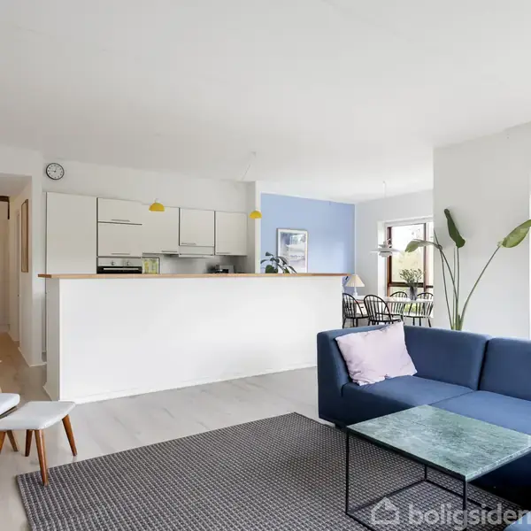 Blå sofa står i moderne stue, foran glasbord. Køkken i baggrunden med hvidt køkkenbord, gule lamper over. Lyse træmøbler og grøn plante skaber hyggelig atmosfære i lys lejlighed.