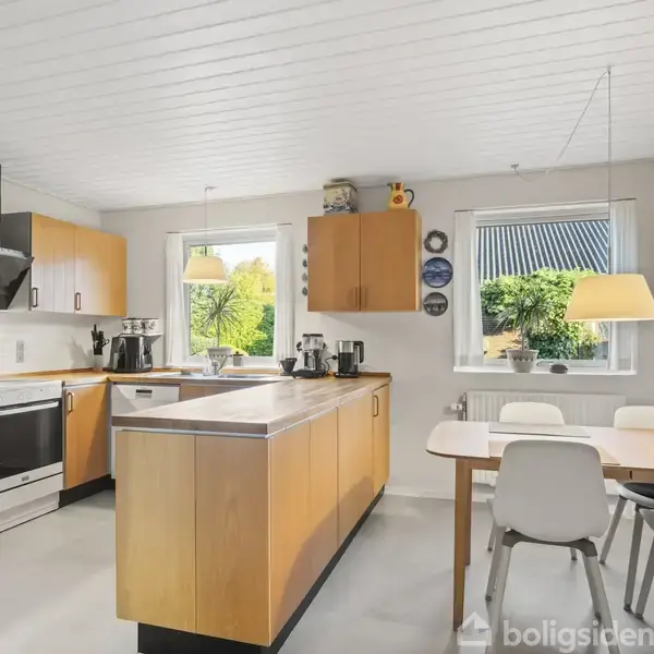 Køkken med træskabe og bordplader, ovn og emhætte. Bordet med fire stole står ved vinduerne, der har udsigt til grønt område. Loftslampe hænger over spisebordet.