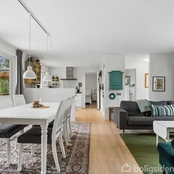 Spisebord med hvide stole står på gulvtæppe, mens sofa og sofabord er placeret på trægulv. Store vinduer til venstre og åben køkken i baggrunden.