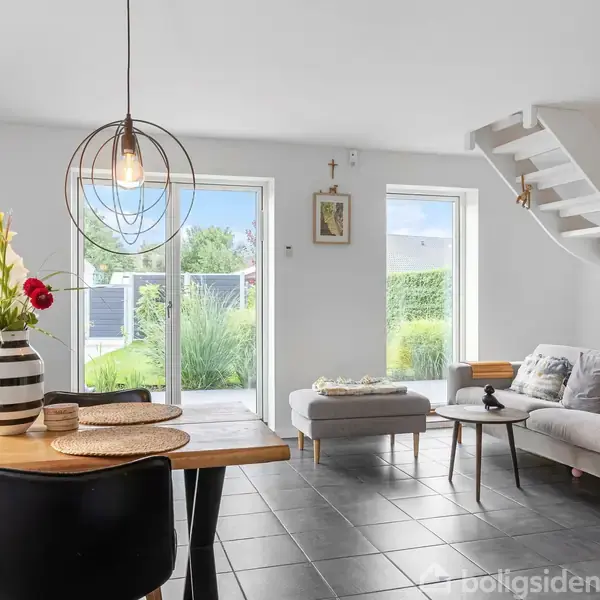 En lys stue med spisebord, der har en vase med blomster. Sofa og sofabord står ved siden af en dør til haven. Trappen fører til første sal.