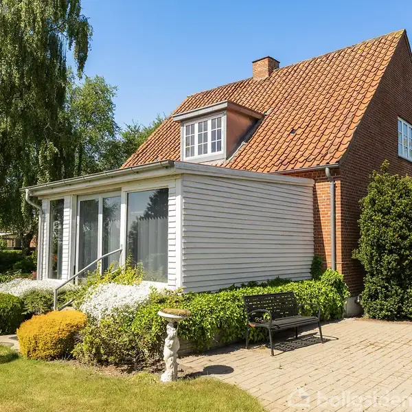 Rødstenshus med skråt tegltag og stor glaseret veranda. Haven har græsplæne, buske og havemøbler på et stenbelagt område. Solskinsvejr skaber en lys og indbydende atmosfære.