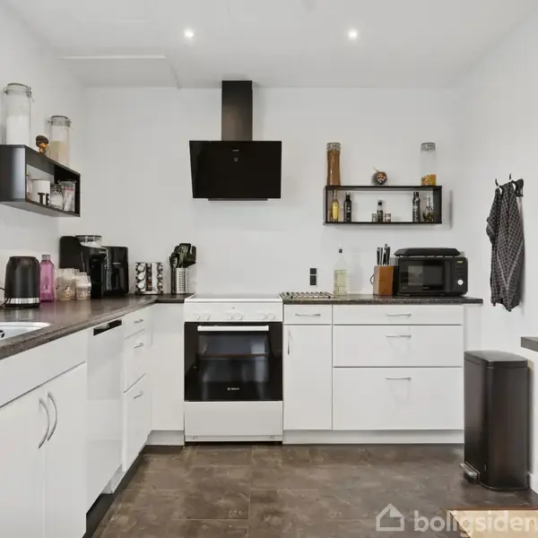 Et hvidt køkken med brune bordplader; der står en ovn med emhætte. Små hylder indeholder krydderier og små krukker. Køkkenapparater og en affaldsspand er placeret langs væggene.