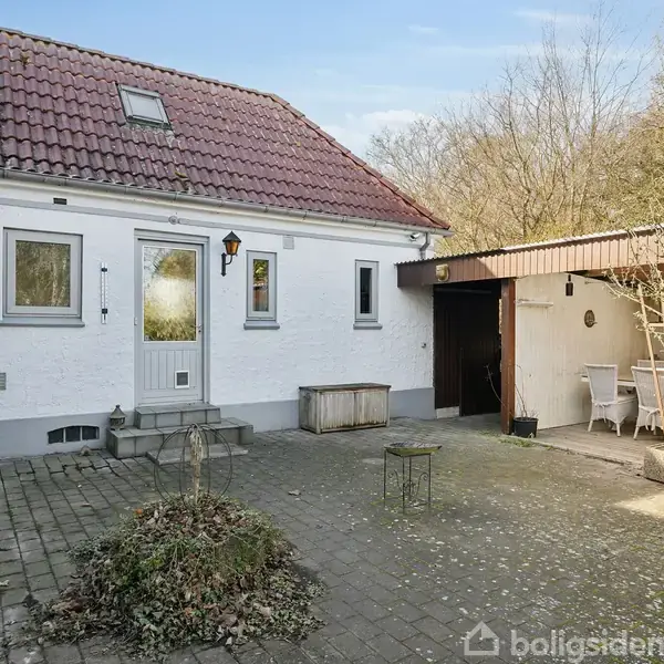 Hvide hus med rødt tegltag og hoveddør, placeret på en flisebelagt gårdsplads. Til venstre er en træbeklædt terrasse med møbler. Omringet af træer og buske, ingen tekst til stede.
