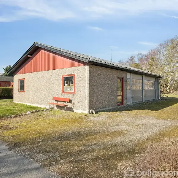 En garagebygning med grå facader og rødt tag ses fra siden, stående på en græsplæne omkranset af træer under en klar blå himmel.