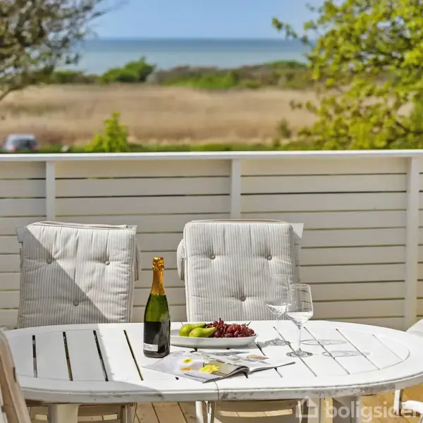 Et rundt bord med hvid dug og stole står på en terrasse. Bordet har en flaske champagne, glas, frugter og et magasin. Baggrunden viser et landskab med marker, træer og havudsigt.