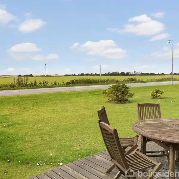Træbord med stole står på en terrasse, omkranset af en grøn græsplæne og en vej i landlige omgivelser med marker og blå himmel med skyer i baggrunden.