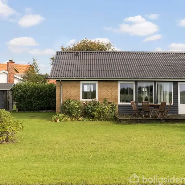 Et lille hus står foran en stor grøn græsplæne med et bord og stole på terrassen. Baggrunden viser flere huse og buske under en blå himmel med skyer.