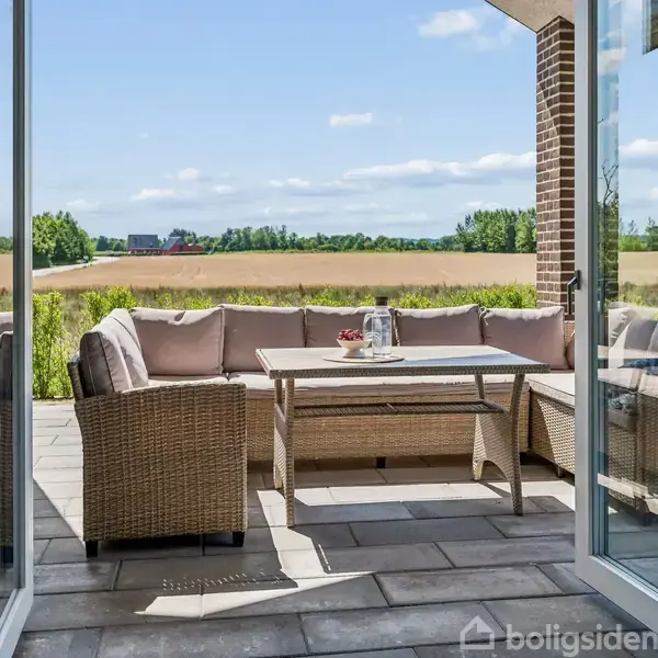 Rattan loungesæt med puder står på en flisebelagt terrasse. En kande og krus står på bordet. I baggrunden ses en mark og en blå himmel med få skyer.