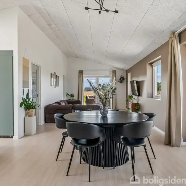 Rundt spisebord med fire sorte stole står tæt på en moderne, åben stue med sofaer. Store vinduer og døre giver udsyn til en terrasse og grønne områder udenfor.