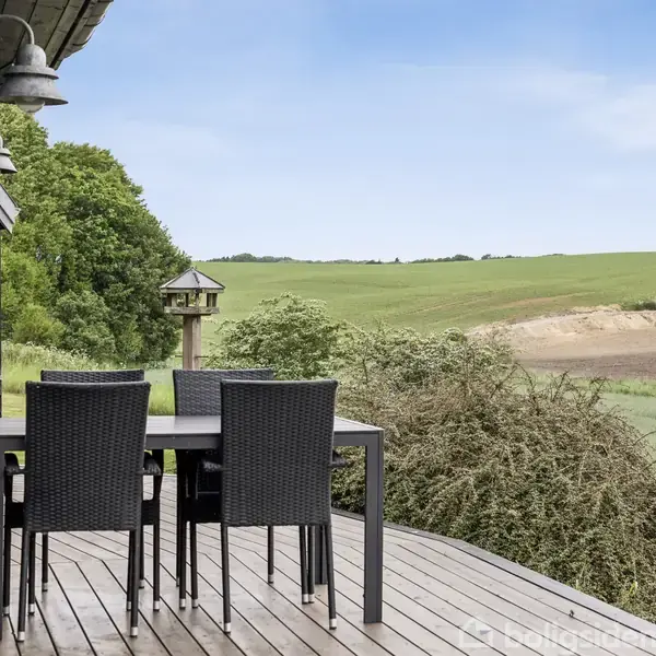 En terrasse med sort bord og stole står udendørs mod en grøn mark med bakker i baggrunden. En vindueskarm og udendørs lamper ses til venstre.