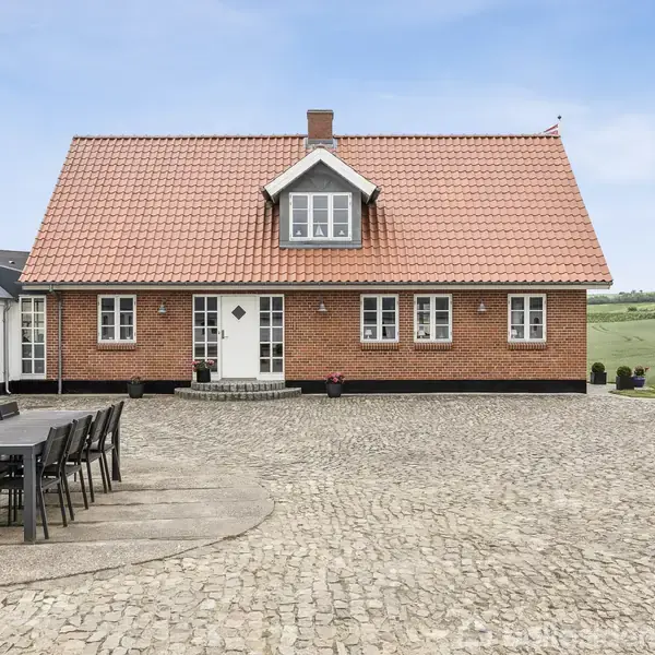 Rødstenshus med rødt tegltag, omgivet af staldbygninger. På den brostensbelagte gårdsplads står et sort havebord med otte stole. I baggrunden ses grønne bakker under en blå himmel.
