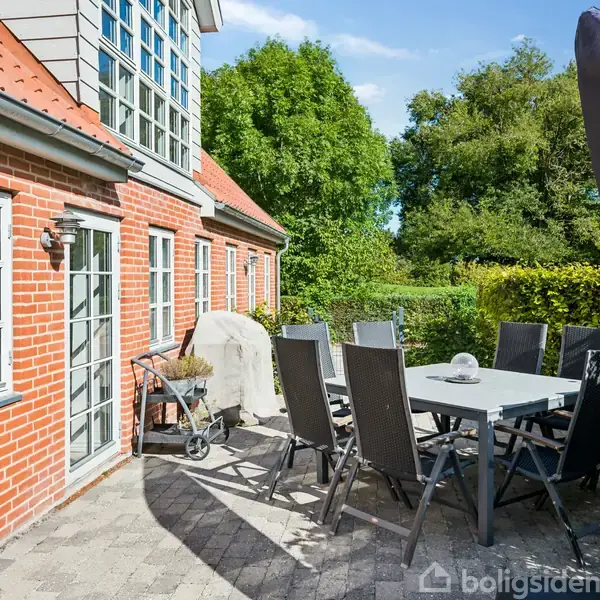 En flisebelagt terrasse foran et murstenshus med bord, stole og grill. Omgivet af grønne buske og træer under en blå himmel. Stor parasol står ved siden af møblerne.