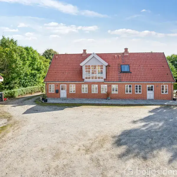 Rødt murstenshus med rødt tegltag, placeret på en grusbelagt gårdsplads. Et køretøj holder til højre ved siden af huset. Omgivet af grønne træer og en klar blå himmel.