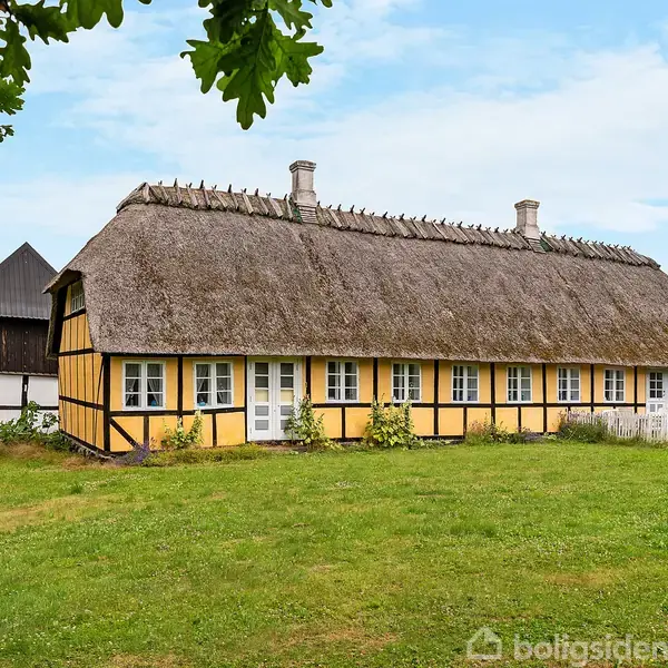 Stråtækt bindingsværkshus står på en grøn eng. Huset har gule vægge med hvide vinduer og ligger i landlige omgivelser med træer, en ubrugt bygning til venstre og en blå himmel.