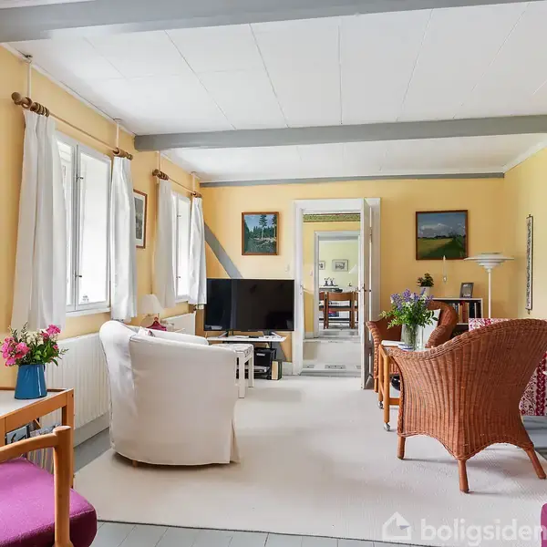 Stue med lysegule vægge og hvide lofter, møbleret med sofaer, stole og et tv. Vinduet er dekoreret med gardiner, omgivet af malerier og blomster i vaser på bordene.