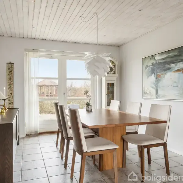 Spisebord med seks stole står i et lyst, moderne rum. Vægkunst og en unik loftslampe tilføjer stil, mens store vinduer slipper naturligt lys ind og viser en udsigt udenfor.