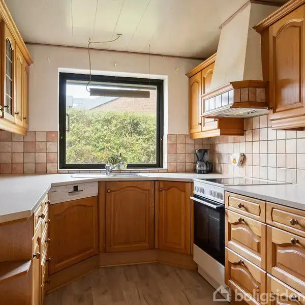 Køkken med træskabe og skuffer arrangeret i en U-form, omkranser en kogeplade og ovn. Fliser pryder væggene, og et vindue giver udsigt til grønne buske udenfor.