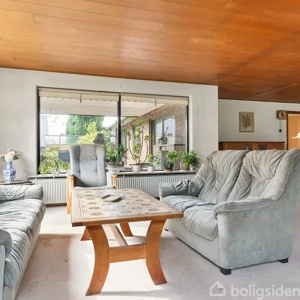 Gråt sofaarrangement står i en lys stue med stort vindue, hvor der ses planter udenfor. Træbord i midten. Lyst trægulv og træloft, med spisestue og lampe i baggrunden.