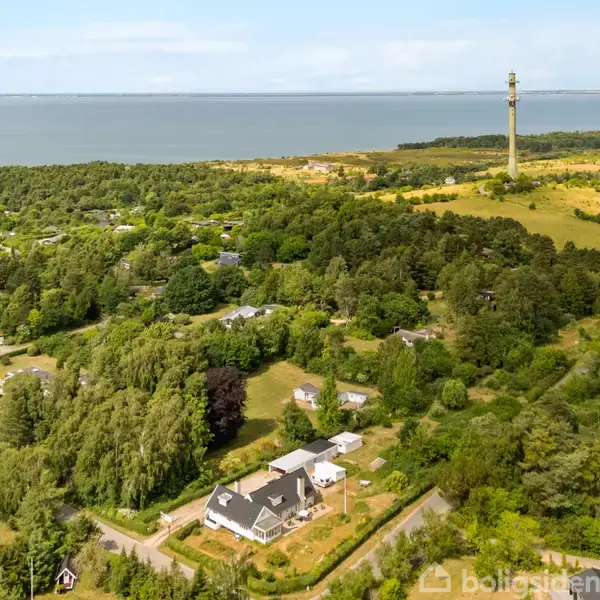 En klynge af huse omgivet af tæt skov, med en høj telekommunikationstower i baggrunden nær en kystlinje. Havet strækker sig til horisonten under en klar himmel.