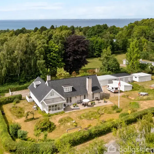 Hus med sort tag står fredeligt omgivet af frodige haver og grønne træer. En flagstang står i forhaven. I baggrunden ses en skov og et glimt af havet.