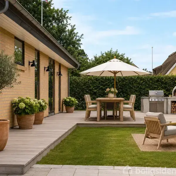 Terrasseområde med trædæk ved et hus med gule mursten. Et rundt havebord med parasol, omringet af stole. Udendørs køkken med pizzaovn i baggrunden. Grønne planter i krukker dekorerer området.