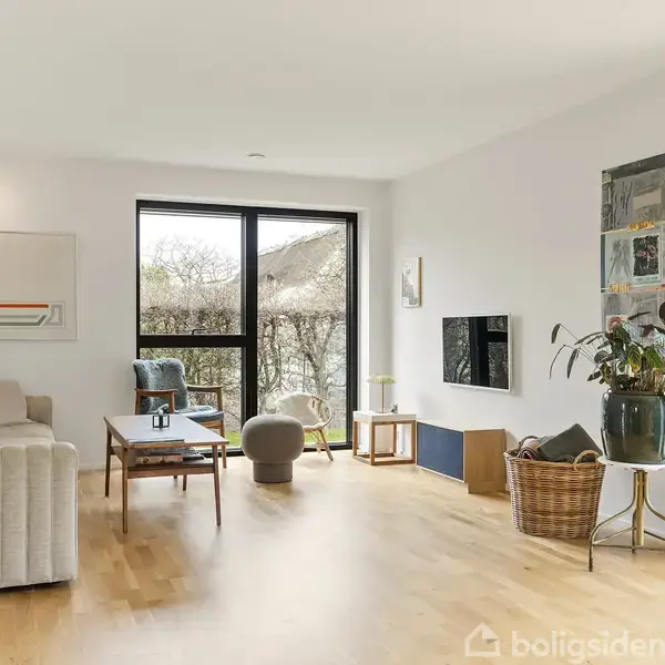 Stue med lyse trægulve har en sofa, sofabord og lænestol. Store vinduer med udsigt til en have skaber lys. Dekorationer og planter kompletterer det moderne, minimalistiske design.