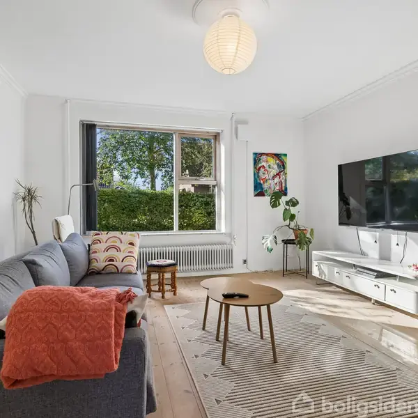 Sofa står ved væggen med en rød plaid, vendt mod et væghængt TV. Et stort vindue giver udsigt til træer. Et sofabord og planter fuldender stuen.