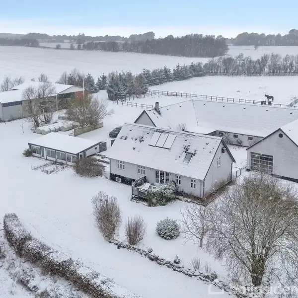 Et landsted dækket af sne står stille om vinteren. Omgivet af sneklædte marker og træer skaber det en idyllisk, fredfyldt landlig scene.