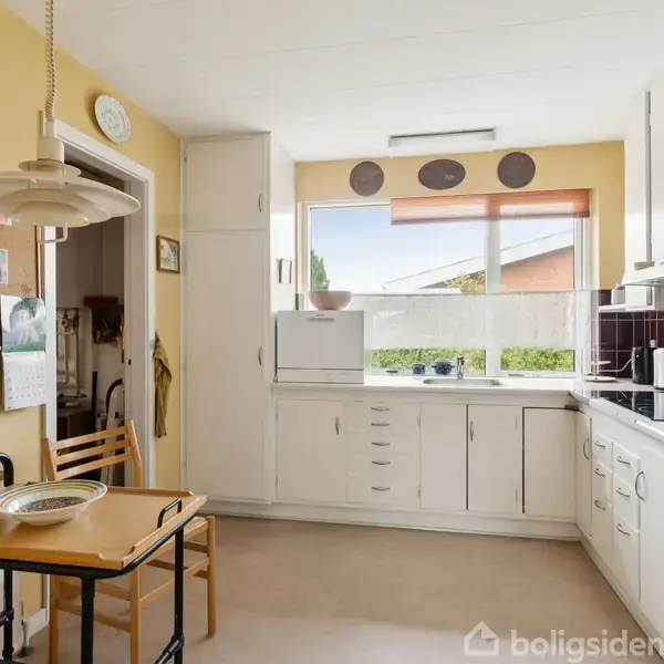 Køkken med hvide skabe, kogeplader og ovn til højre. Bord med stol til venstre. Lys brun væg, stort vindue med udsigt til have. Opslagstavle og ur på væggen.