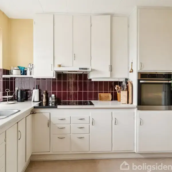 Køkkenet har hvide skabe og skuffer, en vask og en ovn i en lys indstilling. En rødbrun backsplash tilføjer farve. Små apparater står på disken.