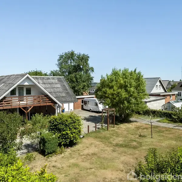 Et hus med stråtag står ved siden af flere andre boliger. Husets terrasse er omgivet af træer og buske i en grøn have. Vejen foran er grusbelagt.