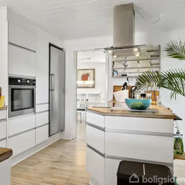 Køkken med hvide skabe og indbyggede apparater. En køkkenø med frugtskål står i midten. En stor plante er placeret ved siden af en glasdør, der leder til spisestuen.