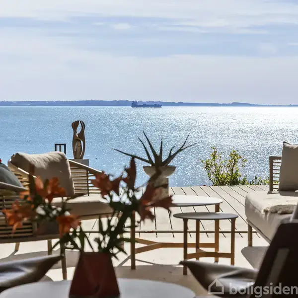 En terrasse med moderne møbler står ud mod en solrig havudsigt. Skibe ses i horisonten, og blomster pryder forgrund.