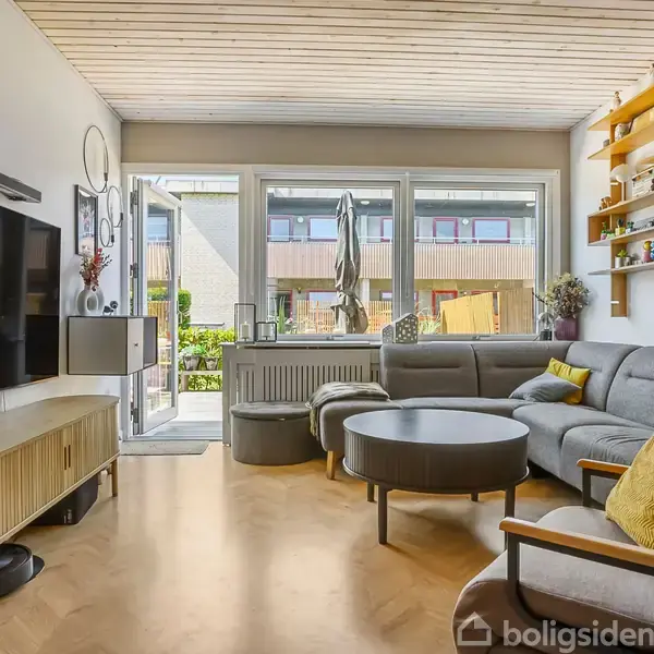 Stue med en hjørnesofa fyldt med farverige puder, placeret foran et rundt sofabord. Væggen har en hylde med dekorationer. Store vinduer åbner ud mod en græsplæne.