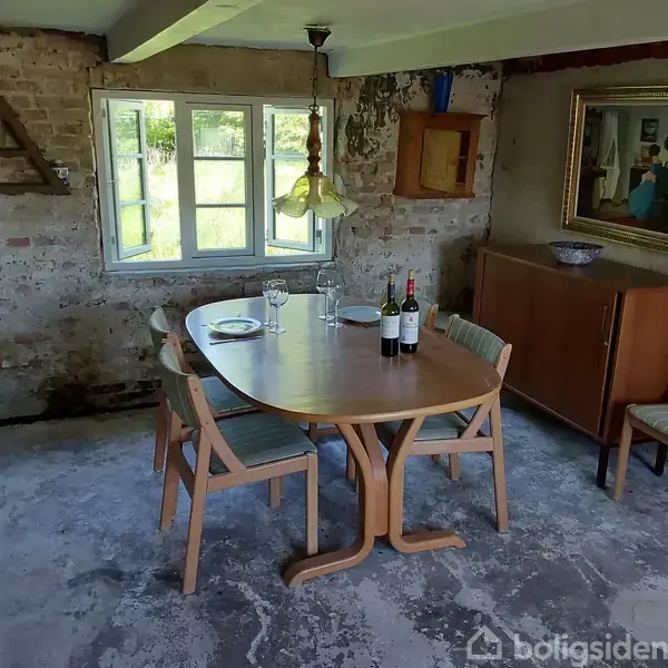 Et træbord med fire stole står i et rum med rå murstensvægge. Ovenpå bordet er der tallerkener, glas og to vinflasker. Vinduet i baggrunden skaber naturligt lys.