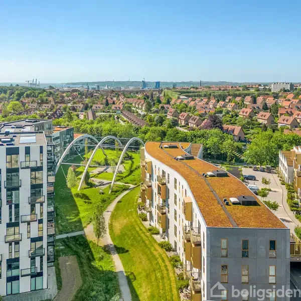 Moderne lejlighedskomplekser med buede tage ses i et grønt byområde. En bro med buer forbinder bygningerne, mens en række huse og skov strækker sig i baggrunden under en blå himmel.