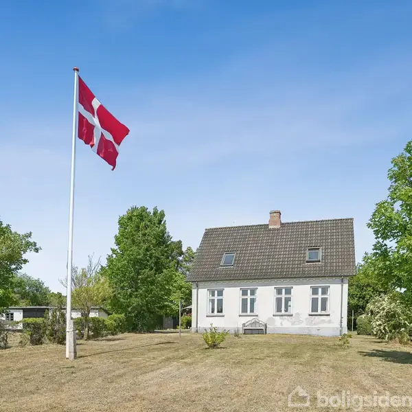Et hvidt hus med mørkt tag står på en stor græsplæne. En dansk flagstang med Dannebrog vejer i vinden. Omkring huset er grønne træer og buske.