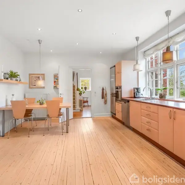 Et lyst køkken med trægulv, hvor et spisebord med stole står til venstre. Store vinduer til højre lader dagslys komme ind. Køkkenet har moderne skabe og apparater.